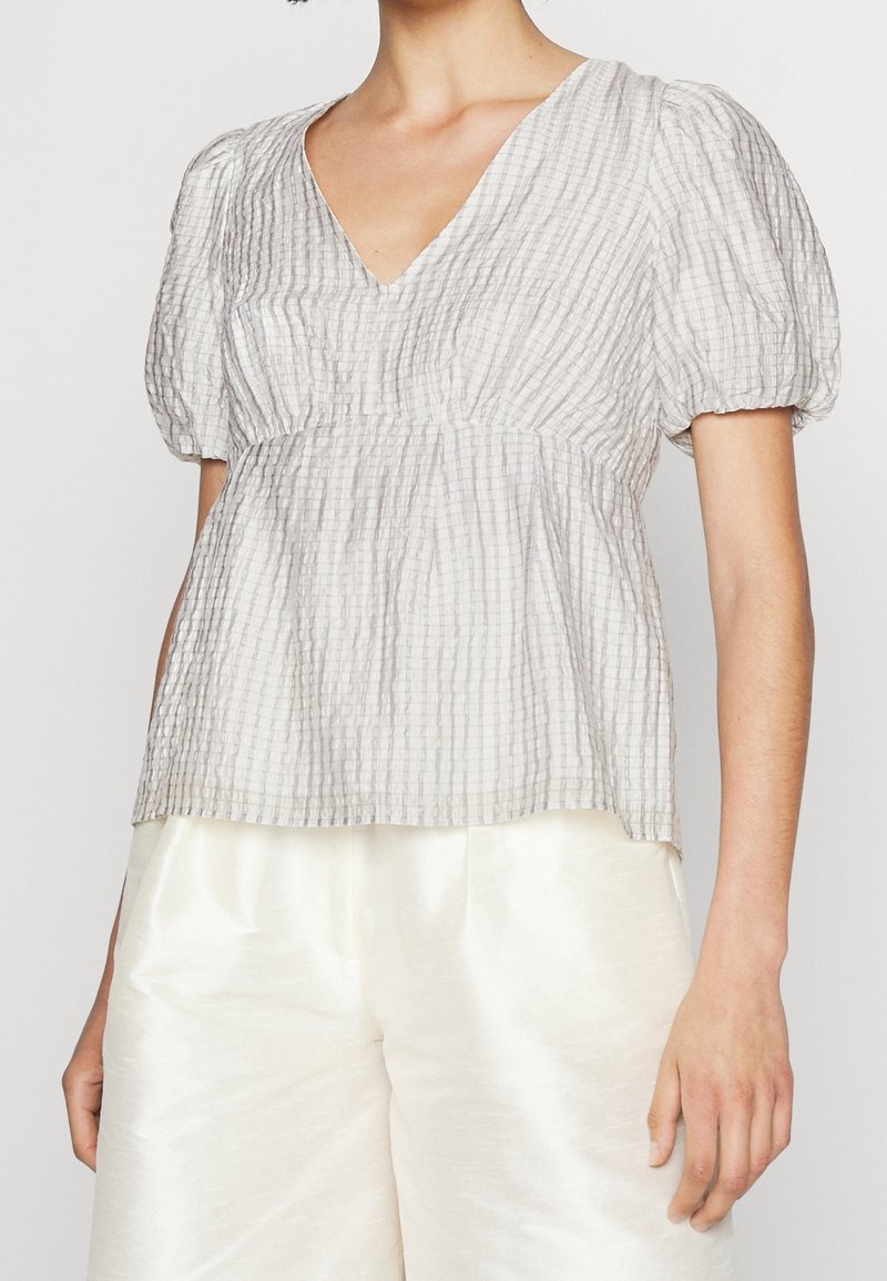 Femme portant un chemisier blanc texturé à manches courtes avec un col en V et une coupe ample, associé à un pantalon large blanc brillant.