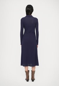 Tmavozelené rebrované šaty s dlhými rukávmi, golierovým výstrihom a opaskom v páse. Dĺžka pod kolená so subtílnymi bočnými rozparkami. Viditeľné hnedé členkové čižmy.