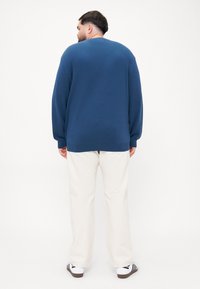Pull en maille bleu à manches longues, coupe décontractée, avec un col côtelé, associé à un pantalon crème et des baskets noir et blanc.