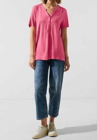 Top corto rosa con maniche corte, scollatura a V e fila di bottoni, abbinato a jeans cropped azzurri e sneakers beige. Texture liscia.