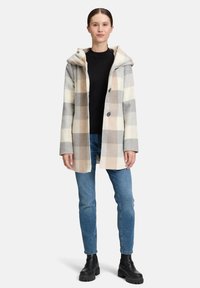 Eine Frau trägt einen beigen, grauen und weißen karierten Mantel über einem schwarzen Oberteil, blauen Jeans und schwarzen Stiefeletten und steht vor einem weißen Hintergrund.