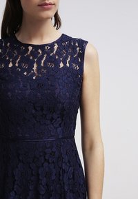 Navy blue lace dress featuring a sheer lace bodice with floral patterns, cap sleeves, and a fitted silhouette, accented by a thin waistband.