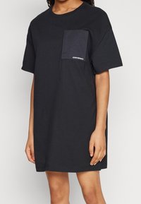Czarna bawełniana sukienka w kształcie t-shirtu z okrągłym dekoltem, krótkimi rękawami oraz jedną kieszenią na piersi z subtelnym akcentem logo.
