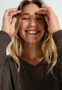 Suéter marrón texturizado con cuello en V, de manga larga. La persona que lo lleva tiene el cabello largo y ondulado rubio y está sonriendo, con las manos en la cara.