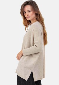 Pull en maille beige à coupe décontractée, avec poignets côtelés et ourlet asymétrique. Texture douce et fentes latérales pour faciliter les mouvements.