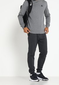 Muž ve šedé mikině, tmavých joggingových kalhotách, černých teniskách a černém batohu Nike stojící na bílém pozadí.