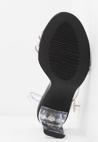 Semelle d'une chaussure à talon noir avec talon en plastique transparent et brides transparentes, vue par le dessous sur un fond blanc.