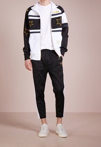 Schwarz-weißes Trainingsanzug mit goldenen Sternakzenten und Streifen, bestehend aus einer Jacke mit Reißverschluss und tapered Hosen. Leichter Stoff. Weiße Sneakers.