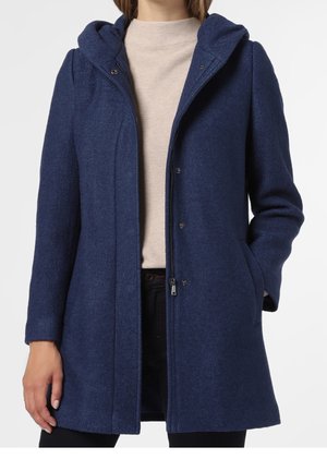 Kvinde iført en mørkeblå hættefrakke over en beige sweater og sorte bukser, med den ene hånd i frakkens lomme og den anden afslappet.