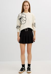 Suéter de punto crema con diseño floral negro, combinado con una falda plisada negra. Zapatillas negras y calcetines de tobillo texturizados complementan el look.