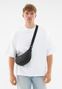 Borsa a tracolla nera realizzata in morbido tessuto, caratterizzata da un design arrotondato, una tracolla regolabile e una piccola patch con logo sul davanti. Indossata a tracolla.