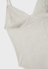 Body en dentelle blanche avec fines bretelles et motifs floraux, avec un buste structuré et des bords délicatement festonnés.