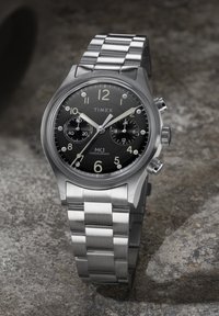 Στρογγυλό ρολόι με μαύρο καντράν Timex MK1 χρονογράφος, με ασημένιο μεταλλικό μπρασελέ και δύο υποκαντράν σε υφασμένη γκρι επιφάνεια.