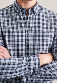 Blå och vit rutig skjorta med knappar, krage, långa ärmar och en ficka; har kontrasterande mörka linjer och texturerad tyg.