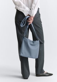 Borsa sintetica blu chiaro con una superficie liscia e logo impresso, portata a spalla; abbinata a pantaloni grigi e ballerine nere.