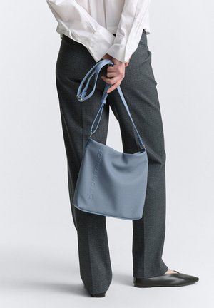 Helleblaue synthetische Handtasche mit glatter Textur und geprägtem Logo, über der Schulter getragen; kombiniert mit grauen Hosen und schwarzen Ballerinas.