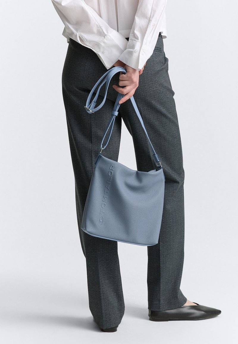 Borsa sintetica blu chiaro con una superficie liscia e logo impresso, portata a spalla; abbinata a pantaloni grigi e ballerine nere.