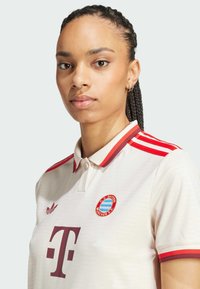 Polo crème avec des accents rouges, doté d'un col boutonné, de trois bandes rouges sur les épaules et du logo du FC Bayern München.