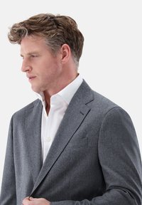 Grau Wollblazer mit schlanker Passform, Reverskragen und einer Brusttasche. Über einem weißen Hemd mit Kragen getragen. Subtile Textur sichtbar.