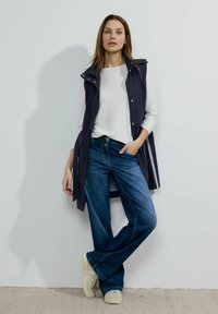 Marinblå lättviktsväst över en vit l�ångärmad topp, i kombination med utsvängda blå jeans och beige sneakers, med en avslappnad passform och dragkedjor.