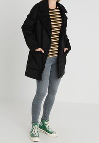 Henkilö, joka käyttää mustaa shearling-takkia sinapinvärisen raidallisen paidan päällä, vaaleanharmaita skinny-farkkuja ja vihreitä korkeita Converse-tennareita, seisoo.