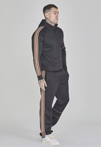 Chándal atlético negro con detalles en rayas naranjas y marrones, sudadera con cremallera y cintura con cordón. Zapatillas blancas completan el look.