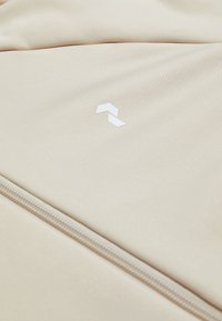 Beige fabric featuring a smooth texture, with a white logo and a zipper detail along one edge.