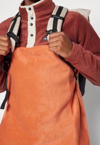 Sudadera de forro polar en color óxido y crema, con un alto cuello, botones a presión y tirantes negros de una mochila.