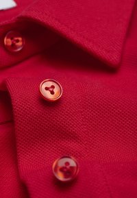 Gros plan sur le tissu d'un polo rouge, présentant un tissage texturé et des boutons rouges brillants à trois trous, mettant en valeur les détails de couleur et de matière.