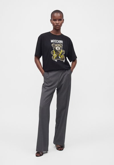 Μοντέλο φοράει μαύρο t-shirt Moschino με γραφικό αρκουδάκι και γκρι παντελόνι φαρδύ με τα χέρια στις τσέπες.