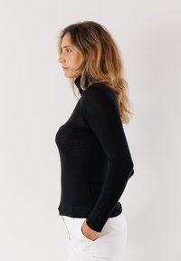 Maglia nera a maniche lunghe con collo alto, design aderente, texture liscia e orlo leggermente curvo, abbinata a pantaloni bianchi.
