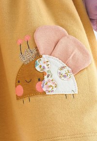 Hérisson brun souriant avec des joues roses, une couronne pailletée, un corps blanc à motifs et de grandes oreilles en tissu rose sur un tissu tricoté moutarde.