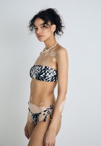 Juodai baltas raštuotas bikini kostiumėlis su bandeau viršumi ir šoninėmis rišamomis kelnaitėmis, pasižymintis lygiu audiniu ir reguliuojamomis petnešėlėmis.