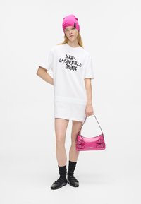 Νεαρή γυναίκα που φοράει λευκό oversized φόρεμα-μπλούζα με κείμενο "Karl Lagerfeld Jeans", φωτεινό ροζ σκουφί, κρατώντας ροζ τσάντα, μαύρες κάλτσες και παπούτσια.