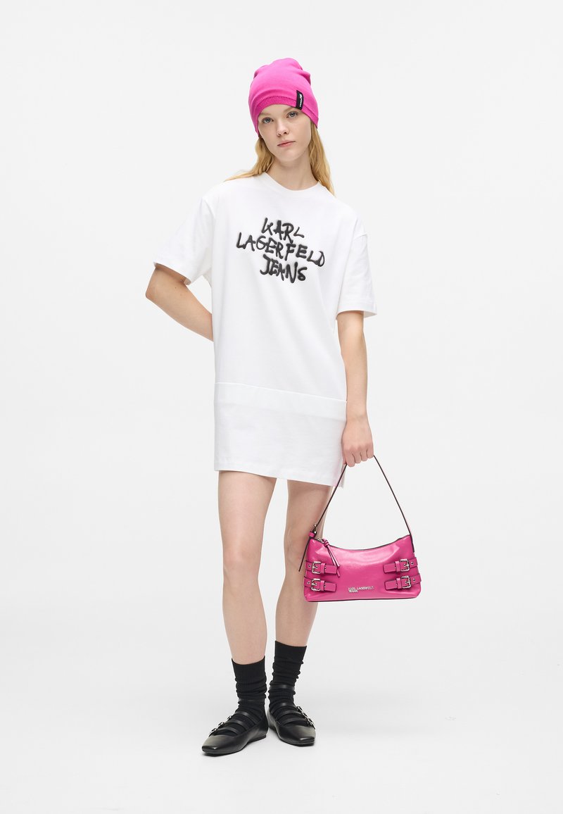 Νεαρή γυναίκα που φοράει λευκό oversized φόρεμα-μπλούζα με κείμενο "Karl Lagerfeld Jeans", φωτεινό ροζ σκουφί, κρατώντας ροζ τσάντα, μαύρες κάλτσες και παπούτσια.