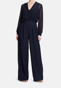 Marineblå jumpsuit med omslagstopp, genomskinliga långa ärmar, vida byxor och plisserade detaljer vid midjan. Slät tygtextur.