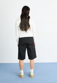 Black shorts with a relaxed fit, paired with a white long-sleeve shirt featuring a circular graphic. Light green sneakers with white accents.