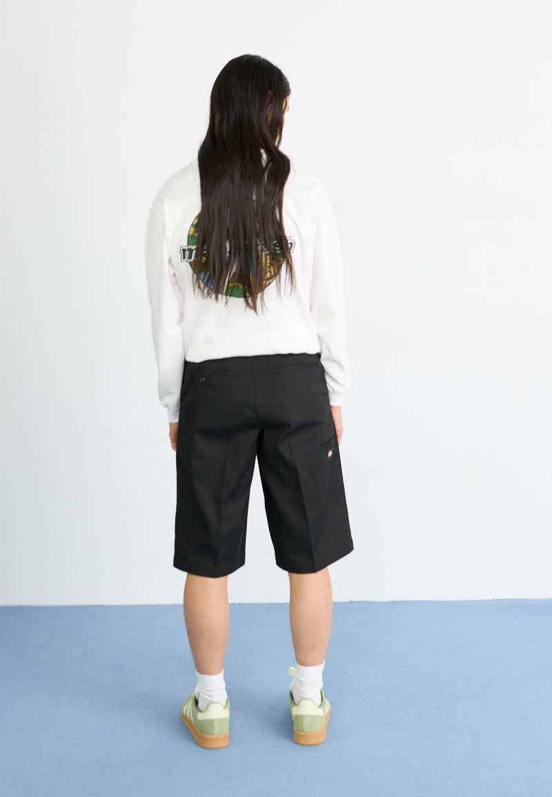 Pantaloni corti neri con vestibilità rilassata, abbinati a una maglietta a maniche lunghe bianca con una grafica circolare. Sneakers verde chiaro con dettagli bianchi.