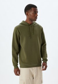 Sudadera verde oliva con capucha ajustable, puños y bajo acanalados, que presenta una textura suave. Llevada con pantalones cargo beiges.
