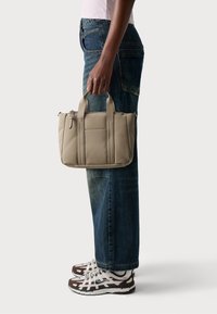 Beige bag with dual top handles and zip closure, featuring stitched accents and a structured design, held beside dark denim jeans and sneakers.