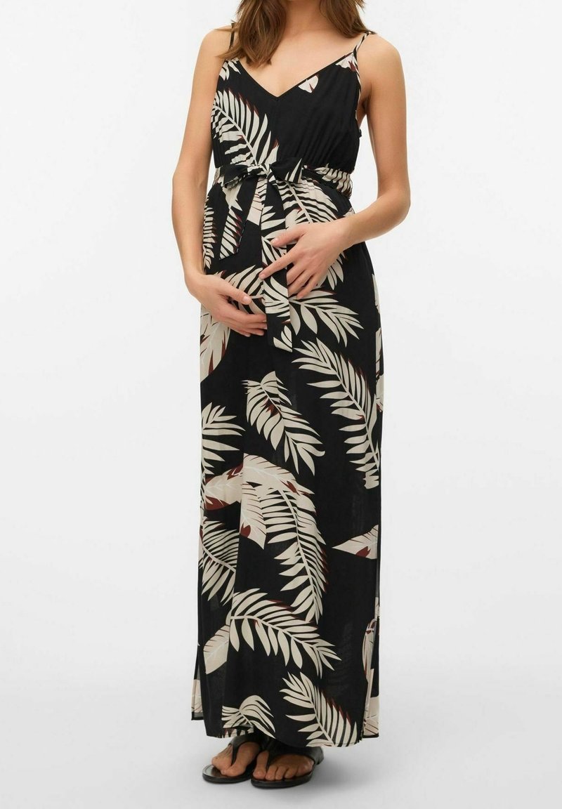 Femme enceinte portant une robe maxi noire avec un motif de feuilles beige, tenant son ventre avec les deux mains, debout pieds nus sur fond blanc.