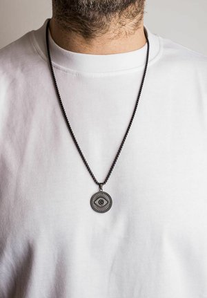 Collana in metallo nero con un pendente circolare che presenta un design a occhio, indossata sopra una maglietta bianca in tessuto testurizzato.