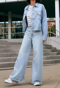 Světle modrá džínsová bunda přes pruhované tričko, spárovaná s volnými světle modrými džínami a bílými teniskami. Hladká textura, neformální design.