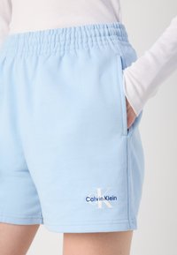 Šviesiai mėlyni sportiniai šortai su elastingu juosmeniu, ant šono išsiuvinėtas Calvin Klein logotipas. Rankos padėtos kišenėje.