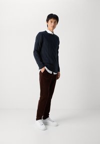 Man som står och bär en marinblå stickad tröja, vit skjorta, bruna byxor och vita sneakers mot en ljus enfärgad bakgrund.