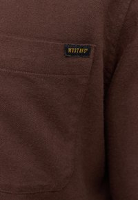 Brauner Stoff mit einer Brusttasche, auf der ein kleines schwarzes Etikett aufgenäht ist, das den gelben Markennamen "MUSTANG" zeigt.