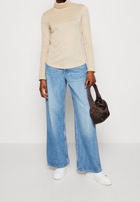 Beige, glinsterende coltrui gecombineerd met lichte blauwe, wijde denim jeans. Houdt een kleine, bruine satijnen handtas vast. Witte sneakers maken de outfit compleet.