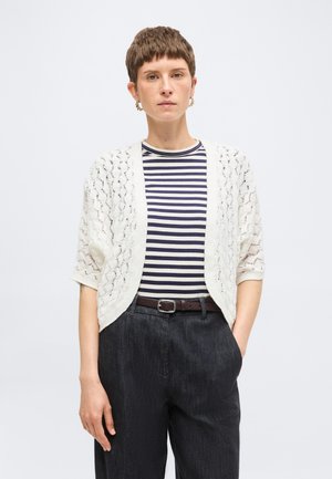 Femme aux cheveux courts portant des boucles d'oreilles créoles en or, un cardigan blanc ajouré, un haut rayé noir et blanc, un pantalon sombre taille haute et une ceinture.