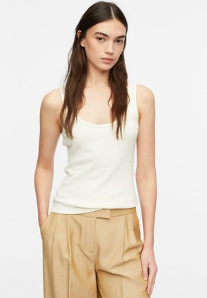 Młoda kobieta o długich brązowych włosach, ubrana w białą bezrękawnik i beżowe spodnie z wysokim stanem, stojąca z rękami w kieszeniach.