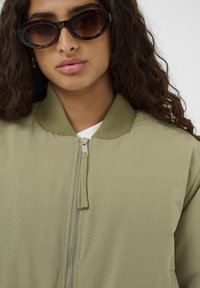 Olive zelena bomberska jakna z teksturiranjem, rebrastim ovratnikom in srebrnim zapiranjem. Črne očala v oklepni obliki z ovalnimi lečami.